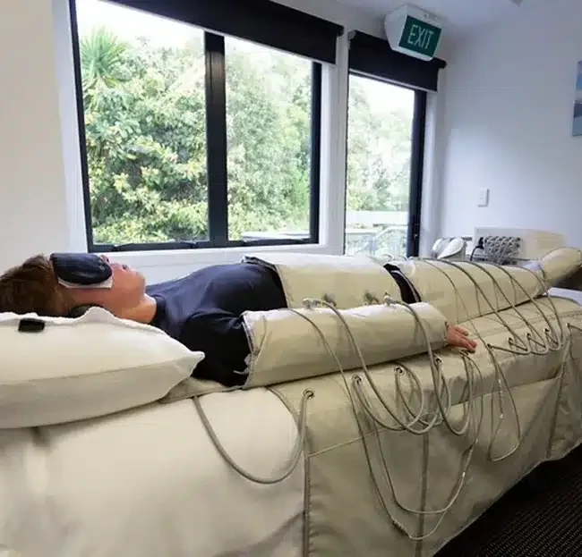 Person lying on a padded medical treatment bed in a clinic, wearing headphones and connected to many cables/sensors.