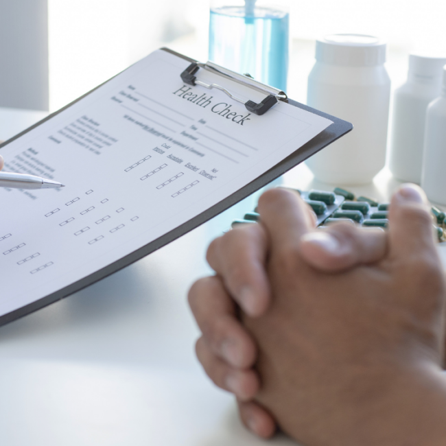 Health check form on a clipboard with a person’s clasped hands in the foreground and medicine bottles on a countertop.