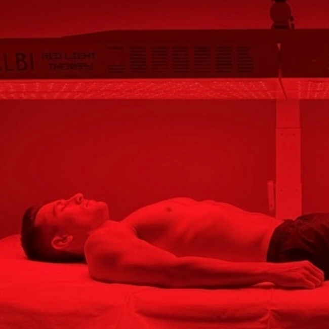 Shirtless man lying on a bed under red light panels, receiving red light therapy treatment.