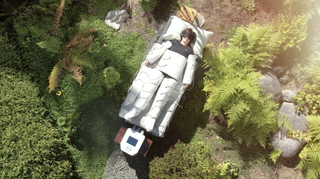 Person lying on a large white padded bed outdoors, viewed from above, with multiple cables attached in a garden setting.