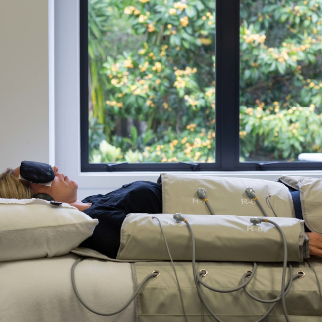 Woman lying on a padded therapy table with multiple electrode pads and wires, wearing a sleep mask near a window.