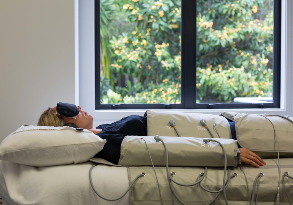 Woman lying on a padded therapy table with multiple electrode pads and wires, wearing a sleep mask near a window.