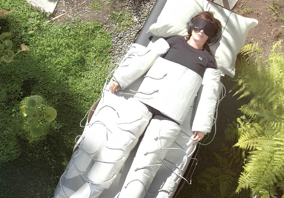Person wearing a sleep mask and headphones resting in a white inflatable medical suit outdoors among plants.