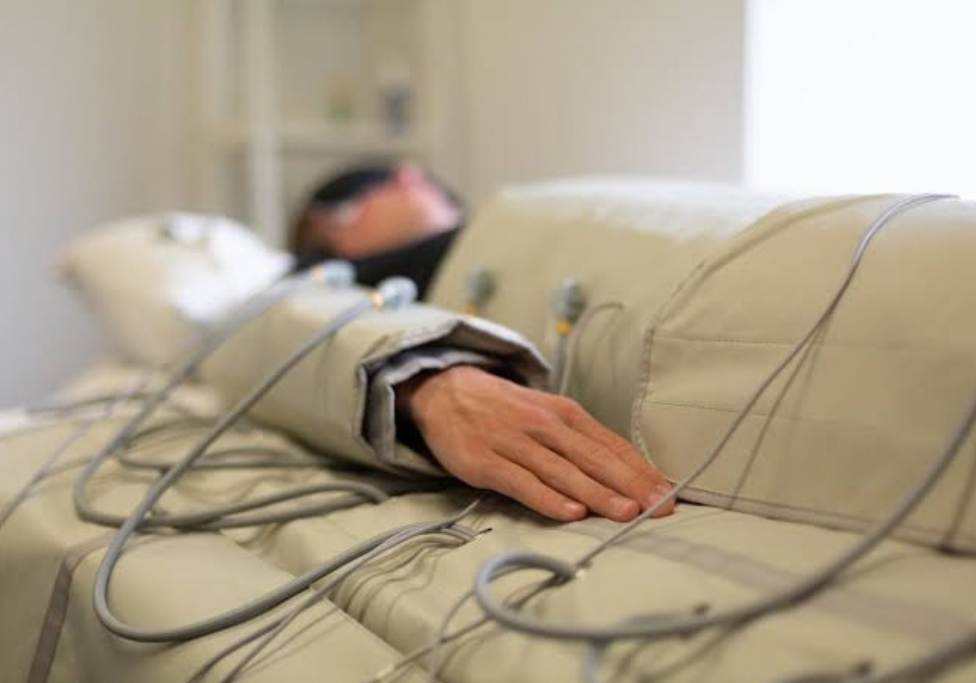 Person resting in a hospital bed with medical cables and sensors attached to their arm and chest area.